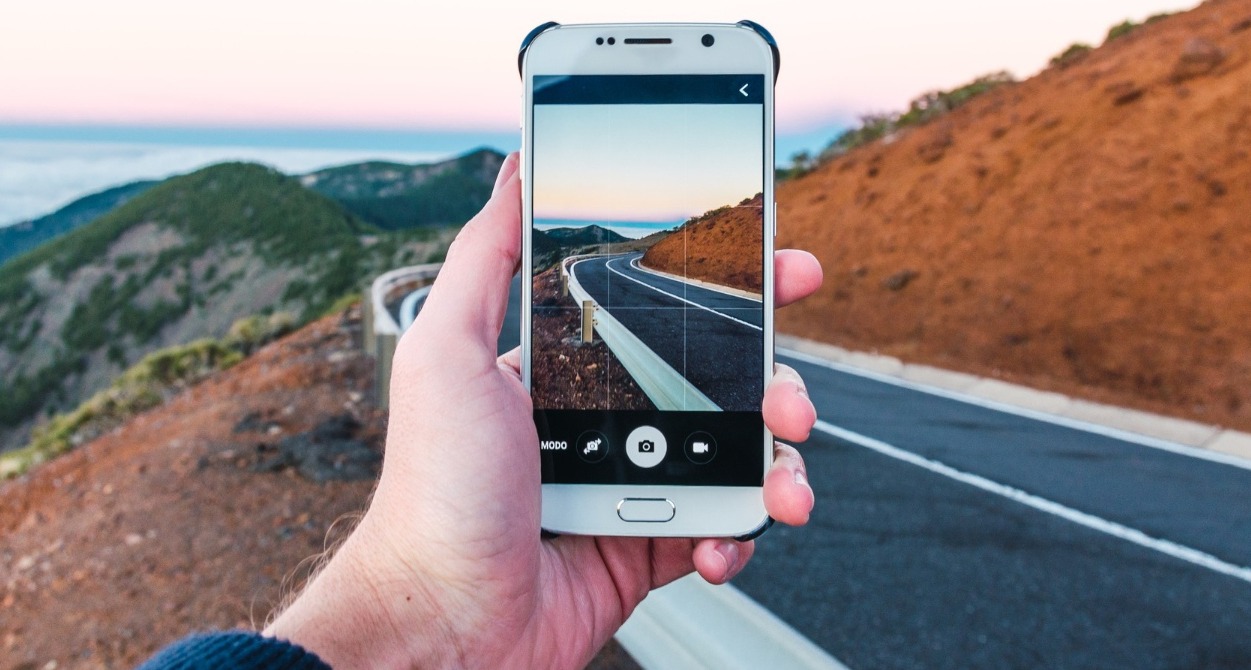 TikTok lanza un nuevo formato publicitario para impulsar reservas en hoteles y destinos turísticos Una mano sosteniendo un móvil para hacer una foto del paisaje de una carretera de montaña
