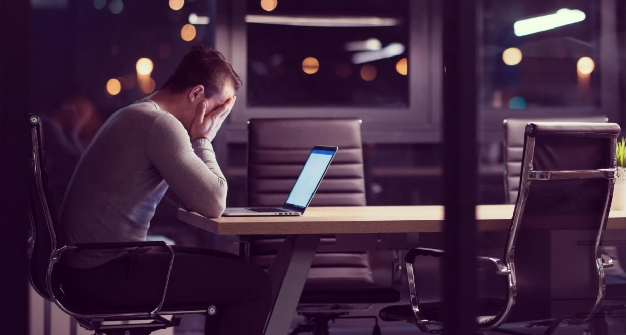 El uso indiscriminado de Inteligencia Artificial en el trabajo está lastrando la productividad, según Stanford un hombre de noche en una sala de reuniones, él solo se echa las manos a la cara en un gesto de desesperación frente al portátil